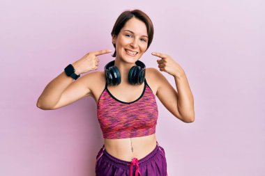 Kısa saçlı, spor giyimli, kulaklık takan neşeli gülümseyen ve parmaklarını ve ağzını gösteren esmer bir kadın. diş sağlığı kavramı. 