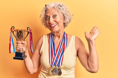 Kıdemli gri saçlı kadın şampiyonluk kupası taşıyor başarıyı mutlu bir gülümsemeyle ve el kaldırarak kutluyor. 