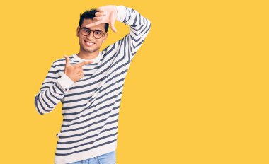 Gündelik kıyafetler ve gözlük takan İspanyol yakışıklı genç adam gülümseyerek elleri ve parmakları gülen yüzlü bir çerçeve yapıyor. yaratıcılık ve fotoğrafçılık kavramı. 