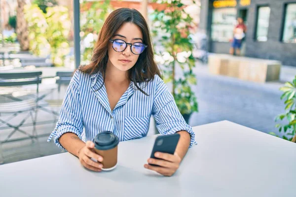 Young hispanic woman with serious expression using smartphone at coffee shop terrace.
