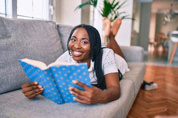 Jovem Afro Americana Sorrindo Feliz Deitado Sofá Leitura Livro Casa — Fotografia de Stock