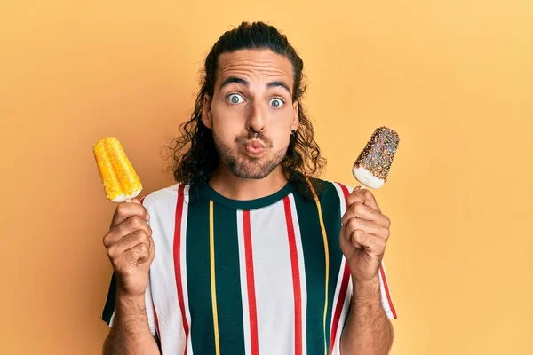 Young handsome man with long hair eating two ice cream puffing cheeks with funny face. mouth inflated with air, catching air.