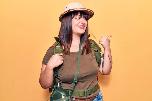 Young plus size woman wearing explorer hat and backpack pointing thumb up to the side smiling happy with open mouth