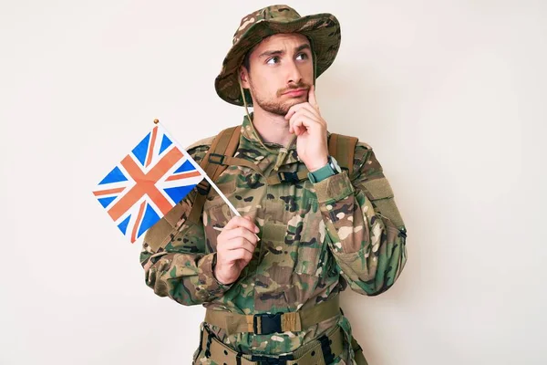 Young caucasian man wearing camouflage army uniform holding united kingdom flag serious face thinking about question with hand on chin, thoughtful about confusing idea