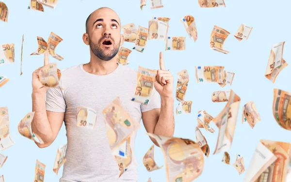 Gündelik tişört giyen genç yakışıklı şaşırmış ve şaşırmış bir şekilde yukarı bakıyor ve parmaklarıyla işaret ediyor ve kollarını kaldırıyor..