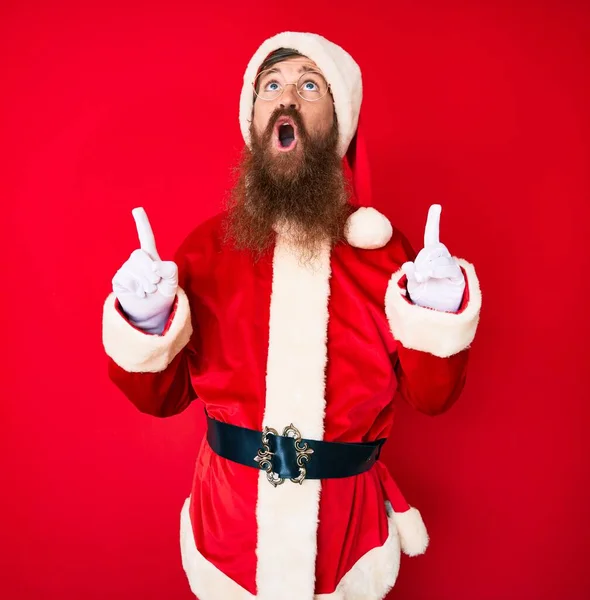 Handsome young red head man with long beard wearing santa claus costume amazed and surprised looking up and pointing with fingers and raised arms.