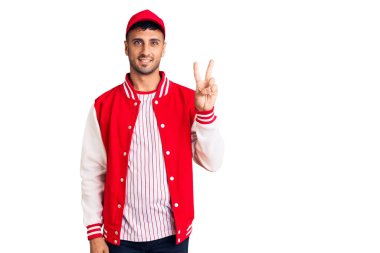 Beyzbol üniforması giyen genç İspanyol adam iki numarayı gösterip işaret ederken kendinden emin ve mutlu bir şekilde gülümsüyor.. 