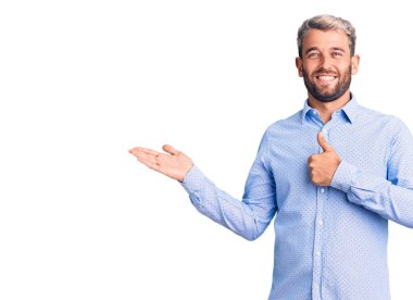 Zarif bir gömlek giymiş genç, sarışın bir adam avuç içini gösteriyor ve baş parmaklarıyla iyi bir jest yapıyor, mutlu ve neşeli gülümsüyor. 