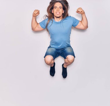 Uzun saçlı, sıradan kıyafetler giyen, mutlu gülümseyen genç yakışıklı adam. Yüzünde tebessümle zıplıyor, beyaz arka planda yumruklar havada kutluyor..