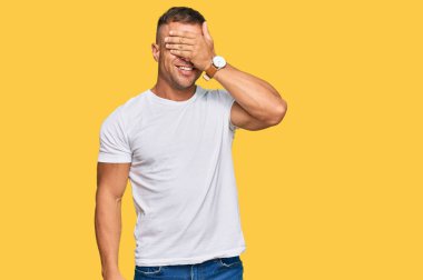 Yakışıklı, sıradan beyaz tişört giyen, gülümseyen ve elleri yüzünde, şaşırtmak için gözleri kapatan kaslı bir adam. kör kavram. 