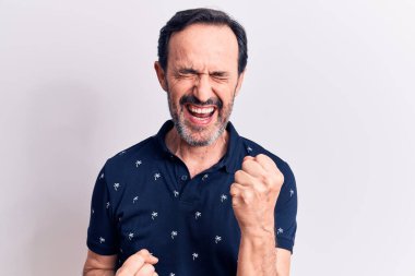 Orta yaşlı yakışıklı bir adam, izole edilmiş beyaz arka planda dikilmiş kolları havada ve gözleri kapalı bir şekilde başarıyı kutluyor ve şaşırıyor.