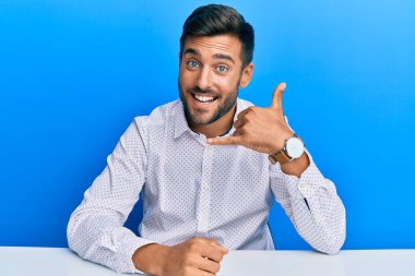 İş kıyafetleri giymiş yakışıklı İspanyol bir adam masaya oturmuş gülümsüyor telefonla el ve parmak hareketleri yapıyor telefonda konuşuyor gibi. iletişim kavramları. 