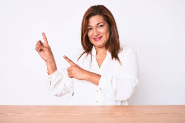 Orta yaşlı esmer, İspanyol iş kadını. Masada oturmuş gülümsüyor ve kameraya bakıyor. İki eliyle ve parmaklarıyla işaret ediyor.. 