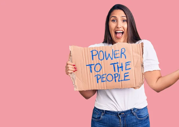 Young beautiful woman holding power to the people cardboard banner celebrating victory with happy smile and winner expression with raised hands