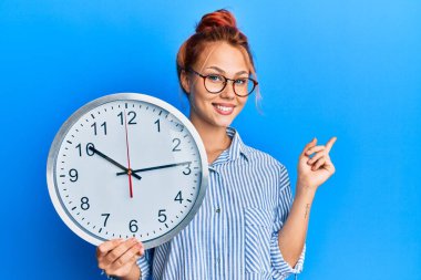 Büyük saati tutan genç kızıl saçlı kadın gülümsüyor mutlu bir şekilde el ve parmak kenarı işaret ediyor. 