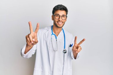 Doktor üniforması ve steteskop giymiş genç yakışıklı adam kameraya bakıp zafer işareti yapıyor. İki numara.. 