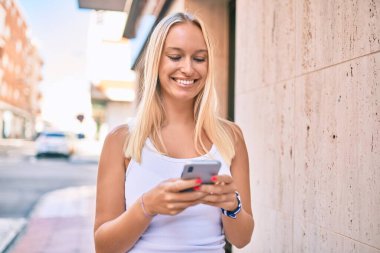 Genç sarışın kız şehirde akıllı telefon kullanarak mutlu bir şekilde gülümsüyor..