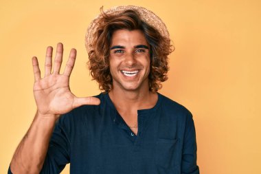 İspanyol asıllı genç adam yaz şapkası takıyor ve beş numarayı gösterip işaret ederken kendinden emin ve mutlu gülümsüyor.. 