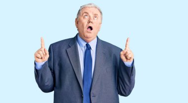 İş ceketi giyen yaşlı gri saçlı adam şaşkın şaşkın yukarı bakıyor ve parmaklarıyla işaret ediyor ve kollarını kaldırıyor.. 