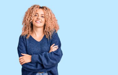 Kıvırcık saçlı, sıradan bir kazak giyen, kollarını kavuşturup kameraya bakan mutlu yüzlü genç sarışın kadın. pozitif kişi. 