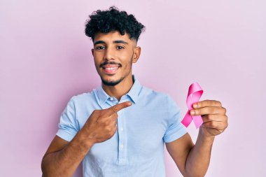 Genç Arap yakışıklı adam pembe kanser kurdelesi tutuyor gülümsüyor mutlu bir şekilde eli ve parmağı ile işaret ediyor 