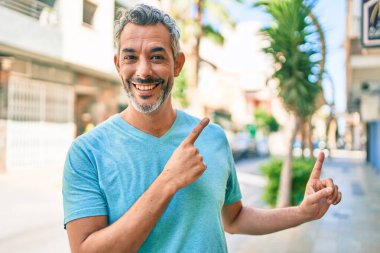Şehrin caddesinde günlük kıyafetler giyen, gülümseyen ve kameraya bakan orta yaşlı, gri saçlı bir adam. İki eliyle ve parmaklarıyla işaret ediyor.. 