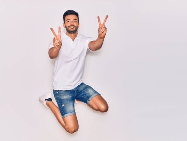 Genç, yakışıklı, sıradan kıyafetler giyen, mutlu gülümseyen bir İspanyol. Yüzünde gülümsemeyle zıplıyor zafer işareti yapıyor parmakları izole beyaz arka planda.