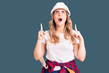 Güvenlik kaskı takan genç beyaz kadın şaşırmış ve şaşırmış bir şekilde yukarı bakıp parmaklarıyla işaret ediyor ve kollarını kaldırıyordu.. 