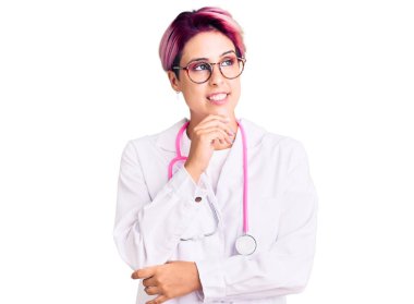 Pembe saçlı, doktor üniforması giyen, elleri çenesinde soru soran, düşünceli bir kadın. Düşünceli bir yüzle gülümsüyor. şüphe kavramı. 