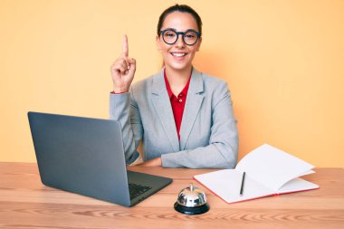 Otel resepsiyonunda çalışan genç esmer kadın bir fikir veya soruyla gülümseyen dizüstü bilgisayar kullanarak mutlu bir yüzle işaret ediyor. 