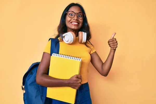 Young indian girl holding student backpack and books smiling happy and positive, thumb up doing excellent and approval sign