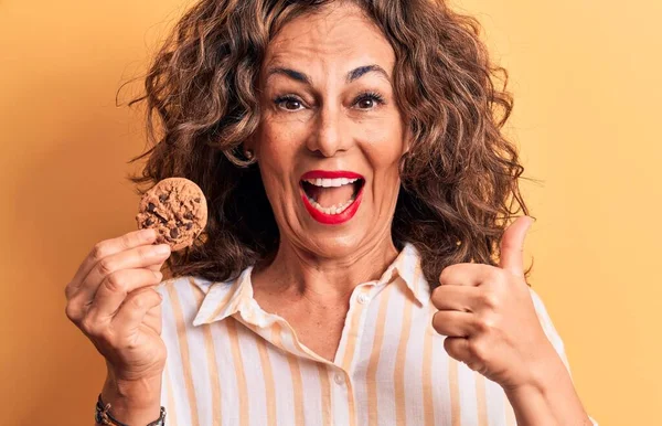 中世ブルネット女性保持Chocoletクッキー上の隔離された黄色の背景Pointing親指へザサイド笑顔幸せとともにオープン口 — ストック写真