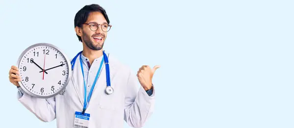 Bonito Homem Hispânico Vestindo Uniforme Médico Segurando Relógio Apontando Polegar — Fotografia de Stock