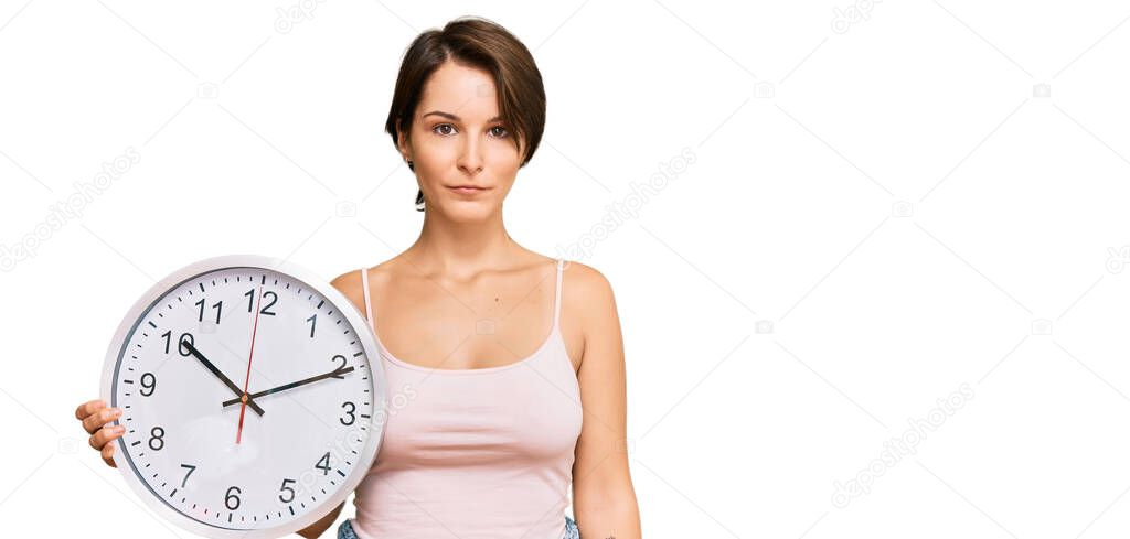 Young brunette woman with short hair holding big clock thinking attitude and sober expression looking self confident 