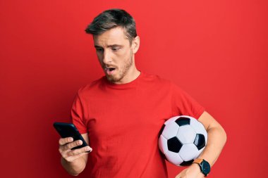 Elinde futbol topu tutan yakışıklı beyaz adam akıllı telefona bakıyor. Şaşırmış ve şaşırmış bir ifade, korku ve heyecanlı bir yüz ifadesi var.. 