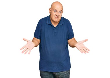 Orta yaşlı, kel, sıradan kıyafetler giyen, elleri ve kolları havada, kafası karışmış bir adam. şüphe kavramı. 