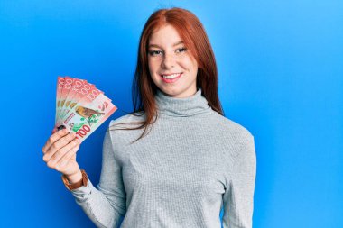 Genç kızıl saçlı kız elinde 100 yeni Zelanda doları banknot tutuyor. Pozitif ve mutlu görünüyor. Kendine güvenen bir gülümsemeyle dişlerini gösteriyor. 