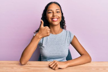 Genç Afro-Amerikan kızı, gündelik giysiler içinde masada oturmuş, elleriyle mutlu parmaklar hareketi yapıyor. Kameraya bakıp başarıyı gösteren ifade. 