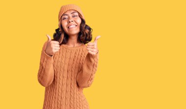 Genç, güzel, melez, yün kazak giyen ve kış şapkalı bir kadın. Elleriyle olumlu jest yapıyor. Gülümsüyor ve mutlu. Neşeli ifade ve kazanan jest. 