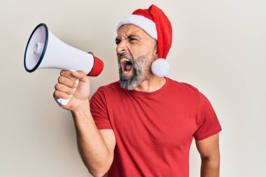 Orta yaşlı, gri saçlı, Noel Baba şapkalı bir adam izole edilmiş beyaz arka plan üzerinde megafon kullanarak bağırıyor..