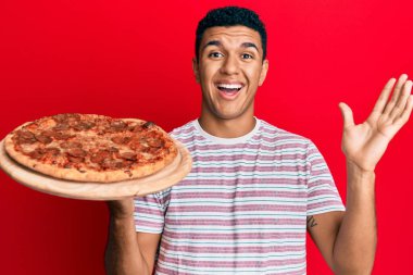 Genç Arap adam İtalyan pizzası tutuyor zaferi mutlu bir gülümsemeyle kutluyor ve kazanan yüz ifadesi kaldırıyor. 