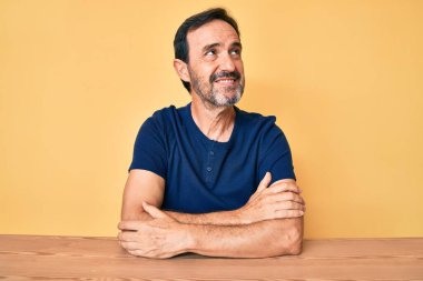 Orta yaşlı İspanyol bir adam, gündelik giysiler giyip masada oturuyor, yüzünde gülümsemeyle yan yana bakıyor, doğal bir ifade. Kendinden emin gülen. 