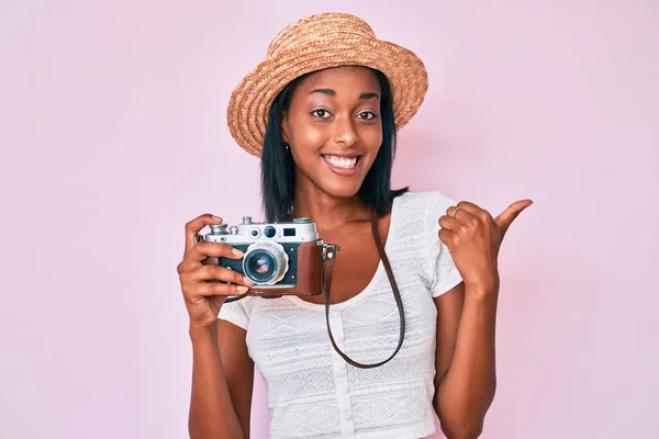 Young african american woman wearing summer hat holding vintage camera pointing to the back behind with hand and thumbs up, smiling confident