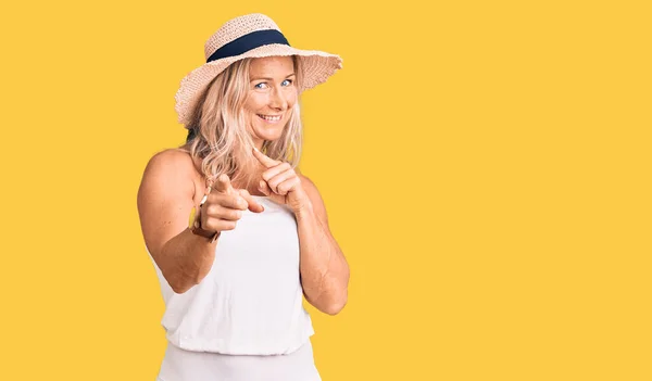 Middle age fit blonde woman wearing summer hat pointing fingers to camera with happy and funny face. good energy and vibes.