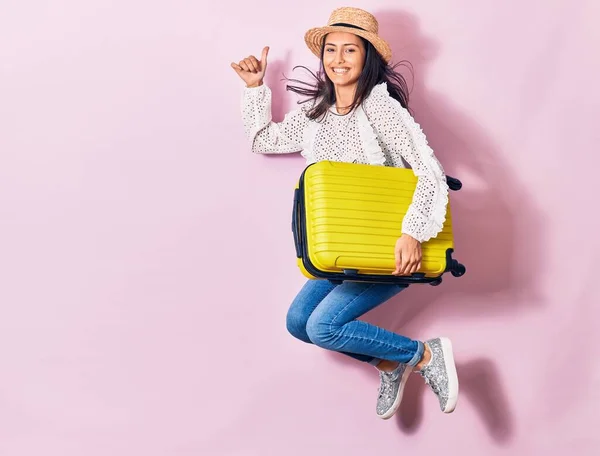 Young beautiful woman on vacation wearing summer clothes and hat smiling happy. Jumping with smile on face holding cabin bag over isolated background