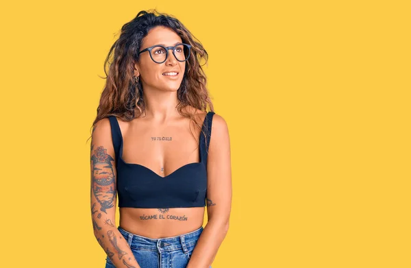 Young hispanic woman with tattoo wearing casual clothes and glasses looking away to side with smile on face, natural expression. laughing confident.