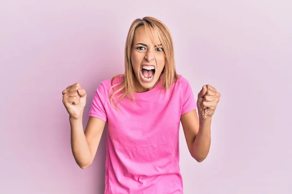 漂亮的金发女人穿着宽松的粉色T恤 怒气冲冲地举起拳头 怒气冲冲地大喊大叫 愤怒和好斗的概念 — 图库照片