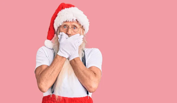 Old senior man with grey hair and long beard wearing santa claus costume with suspenders shocked covering mouth with hands for mistake. secret concept.