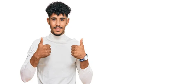 年轻的阿拉伯男子穿着宽松的冬季毛衣 成功地做了一个积极的手势 竖起大拇指 笑容满面 快乐无比 快乐的表达和胜利的姿态 — 图库照片