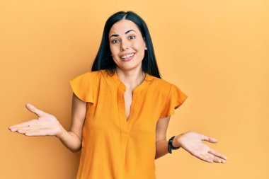 Gündelik kıyafetler giyen, elleri ve kolları havada, kafası karışmış güzel genç bir kadın. şüphe kavramı. 